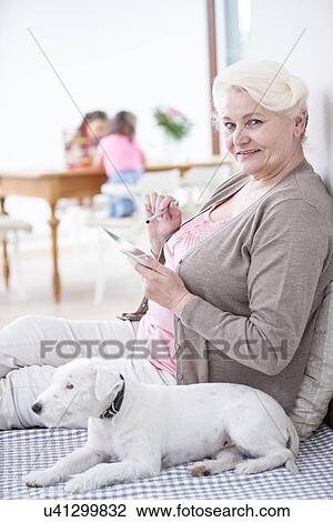 Stock Image - Portrait of happy senior woman using digital tablet by dog at home. Fotosearch
