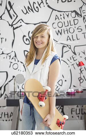 Stock Image - Portrait of happy teenage girl holding skateboard at home. Fotosearch