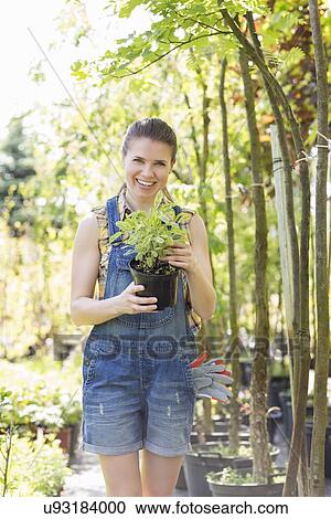 Stock Image - Portrait of happy woman holding potted plant at garden. Fotosearch