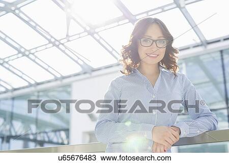 Stock Image - Portrait of happy young businesswoman leaning on railing in office. Fotosearch