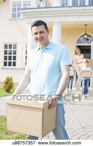 Stock Photo - Portrait of man carrying cardboard box while moving house with family in background. Fotosearch