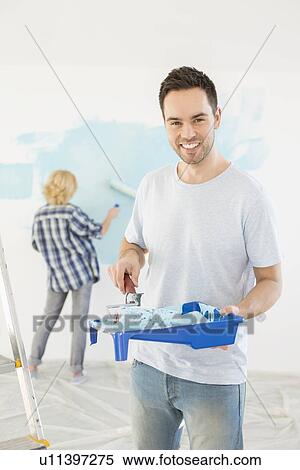 Stock Photography - Portrait of man holding paint roller and tray with woman painting wall in background. Fotosearch