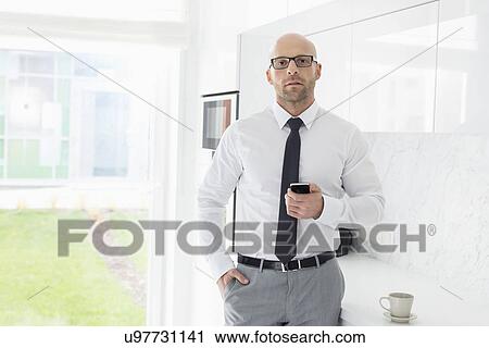Stock Image - Portrait of mid adult businessman holding smart phone at home. Fotosearch