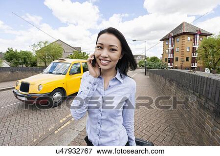 Stock Photo - Portrait of smiling businesswoman answering cell phone on sidewalk with taxi in background. Fotosearch