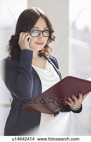 Picture - Portrait of smiling businesswoman using cell phone while holding file in office. Fotosearch