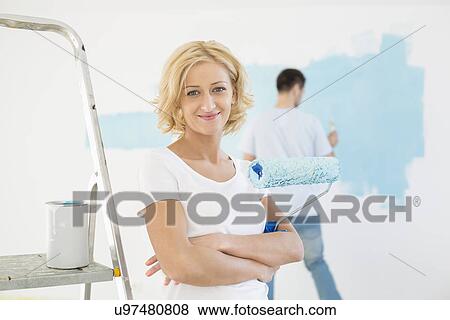 Stock Photo - Portrait of woman holding paint roller with man painting wall in background. Fotosearch