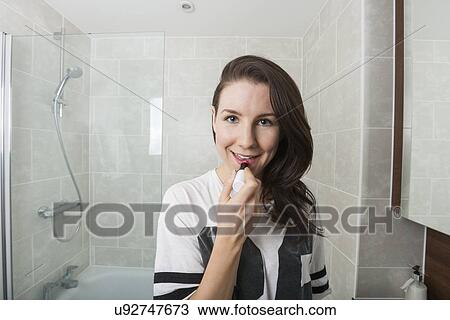 Stock Image - Portrait of young woman applying lipstick in bathroom. Fotosearch