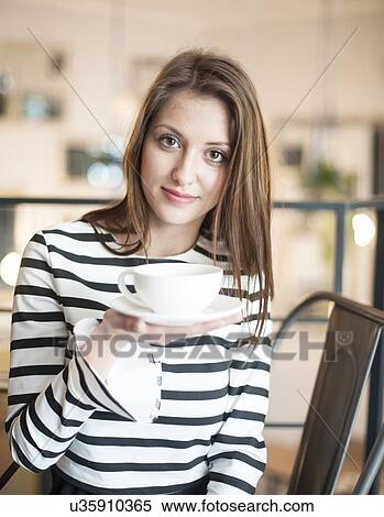 Stock Photography - Portrait of young woman holding coffee cup and saucer at cafe. Fotosearch