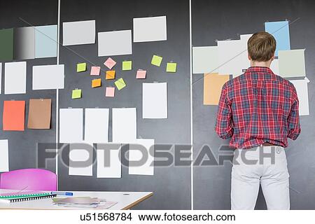 Picture - Rear view of businessman analyzing documents on wall at creative office. Fotosearch