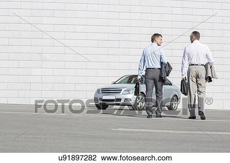 Stock Image - Rear view of businessmen carrying briefcases while walking towards car on street. Fotosearch