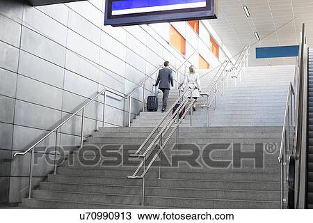 Stock Image - Rear view of businesspeople with luggage moving upstairs in railroad station. Fotosearch