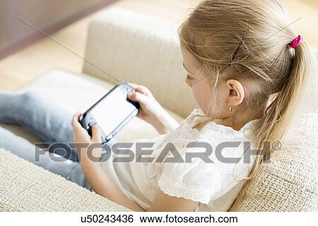 Stock Photograph - Rear view of girl playing hand-held video game at home. Fotosearch