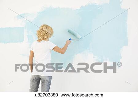 Stock Image - Rear view of woman painting wall with paint roller. Fotosearch
