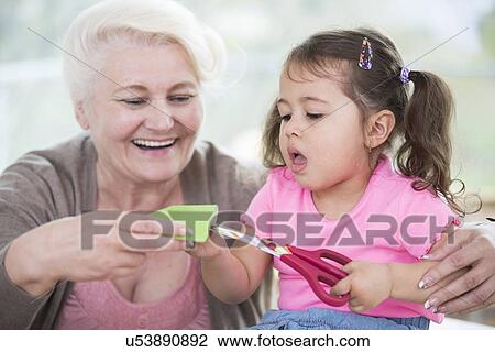Stock Image - Senior woman with granddaughter cutting paper at home. Fotosearch