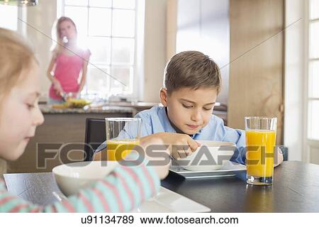 Stock Photo - Siblings having breakfast at table with mother preparing food in background. Fotosearch