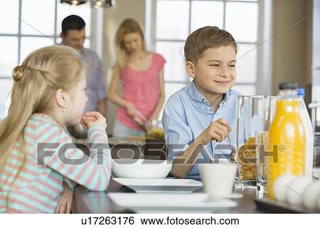 Siblings having breakfast at table with parents cooking in background View Large Photo Image Stock Photograph - Siblings having breakfast at table with parents cooking in background. Fotosearch