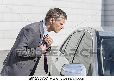 Side view of businessman adjusting tie while looking in car window View Large Photo Image Stock Photo - Side view of businessman adjusting tie while looking in car window. Fotosearch