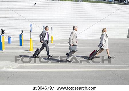 Stock Photo - Side view of businesspeople with luggage walking on street. Fotosearch
