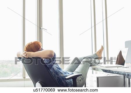 Stock Photo - Side view of businesswoman relaxing with feet up at desk in creative office. Fotosearch