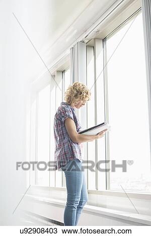Stock Image - Side view of creative businesswoman reading file by window in office. Fotosearch