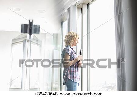 Stock Photo - Side view of creative businesswoman holding files while looking through window in office. Fotosearch
