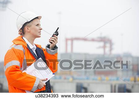 Stock Image - Side view of female engineer using walkie-talkie in shipping yard. Fotosearch