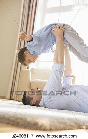 Stock Photo - Side view of playful father picking up son while lying on floor at home. Fotosearch