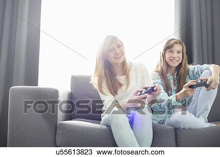 Stock Image - Sisters playing video games on sofa. Fotosearch