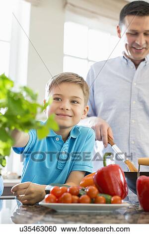 Stock Image - Smiling boy touching houseplant with father preparing food in kitchen. Fotosearch