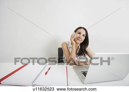 Stock Image - Smiling businesswoman looking away while answering cell phone at desk in office. Fotosearch