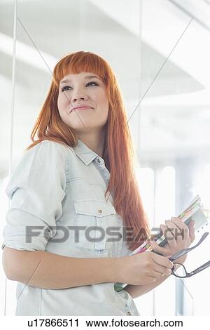 Stock Image - Smiling businesswoman looking away while holding files in creative office. Fotosearch