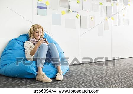 Smiling businesswoman using mobile phone on beanbag chair in creative office View Large Photo Image Picture - Smiling businesswoman using mobile phone on beanbag chair in creative office. Fotosearch