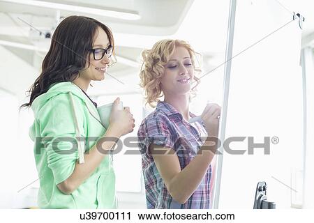 Stock Image - Smiling businesswomen preparing presentation on whiteboard in creative office. Fotosearch