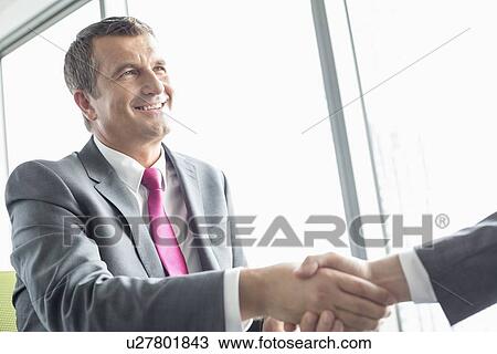 Stock Image - Smiling mature businessman shaking hands with partner in office. Fotosearch
