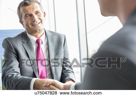 Smiling mature businessman shaking hands with partner in office View Large Photo Image Stock Photo - Smiling mature businessman shaking hands with partner in office. Fotosearch