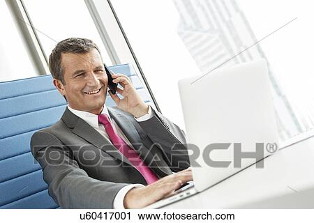 Stock Image - Smiling mature businessman talking on cell phone while using laptop in office. Fotosearch