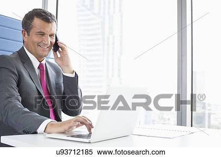 Smiling mature businessman talking on cell phone while using laptop in office View Large Photo Image Stock Photography - Smiling mature businessman talking on cell phone while using laptop in office. Fotosearch