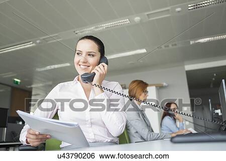 Smiling young businesswoman holding documents while using landline phone with colleagues in background at office View Large Photo Image Stock Photograph - Smiling young businesswoman holding documents while using landline phone with colleagues in background at office. Fotosearch