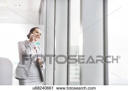 Stock Image - Smiling young businesswoman talking on cell phone near office window. Fotosearch
