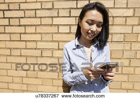 Stock Photo - Smiling young businesswoman text messaging on cell phone against brick wall. Fotosearch