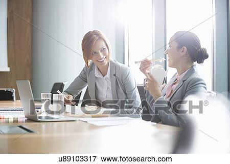 Stock Photo - Smiling young businesswomen having lunch at table in office. Fotosearch