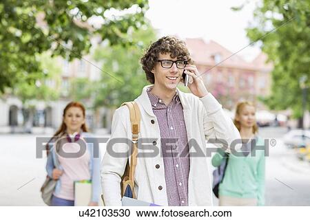 Smiling young male student using cell phone with friends in background on street View Large Photo Image Stock Image - Smiling young male student using cell phone with friends in background on street. Fotosearch