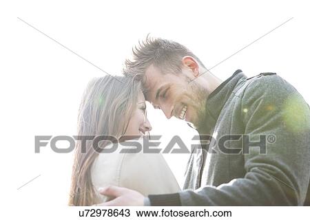Stock Image - Smiling young man and woman embracing against clear sky. Fotosearch