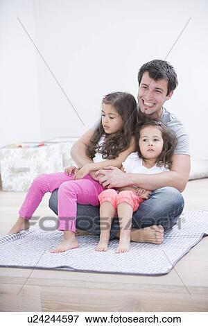 Smiling young man with daughters sitting in his lap at home View Large Photo Image Stock Photo - Smiling young man with daughters sitting in his lap at home. Fotosearch