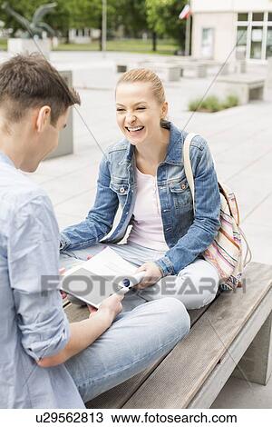 Stock Image - Smiling young woman with male friend studying on bench at college campus. Fotosearch