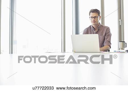 Stock Image - Thoughtful businessman looking away while using laptop at desk in creative office. Fotosearch