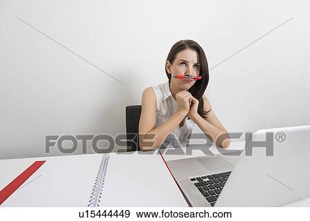 Thoughtful businesswoman holding pen under nose at desk in office View Large Photo Image Stock Photo - Thoughtful businesswoman holding pen under nose at desk in office. Fotosearch