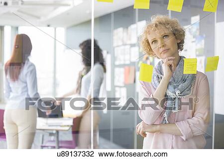 Thoughtful businesswoman reading sticky notes on glass with colleagues in background View Large Photo Image Stock Image - Thoughtful businesswoman reading sticky notes on glass with colleagues in background. Fotosearch