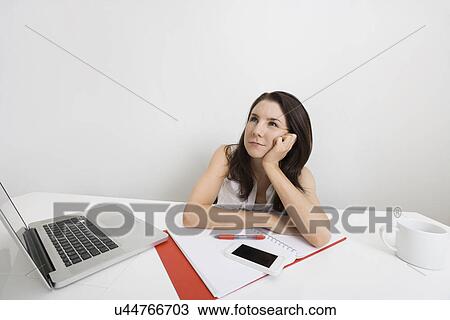 Stock Image - Thoughtful businesswoman with laptop and binder at desk in office. Fotosearch