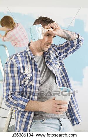 Picture - Tired man holding paint can and brush with woman painting in background. Fotosearch